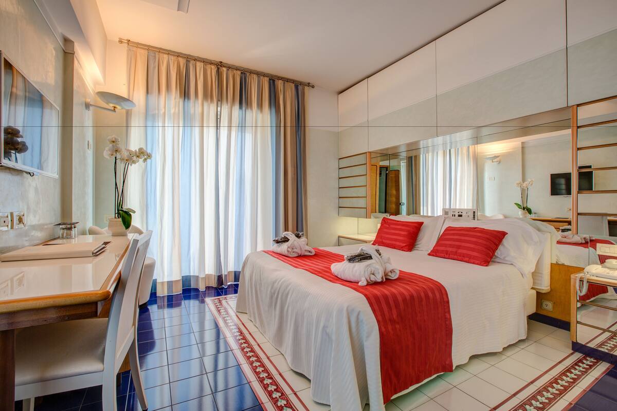 A hotel room with a bed, a desk, a chair, a mirror, and a window with curtains.