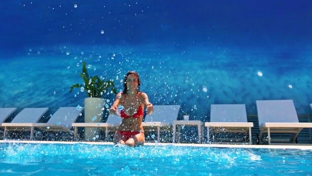 A woman in a red bikini in a pool with water splashing around her. There are lounge chairs and a potted plant in the background. - Atlantic Hotel & SPA (Riccione)