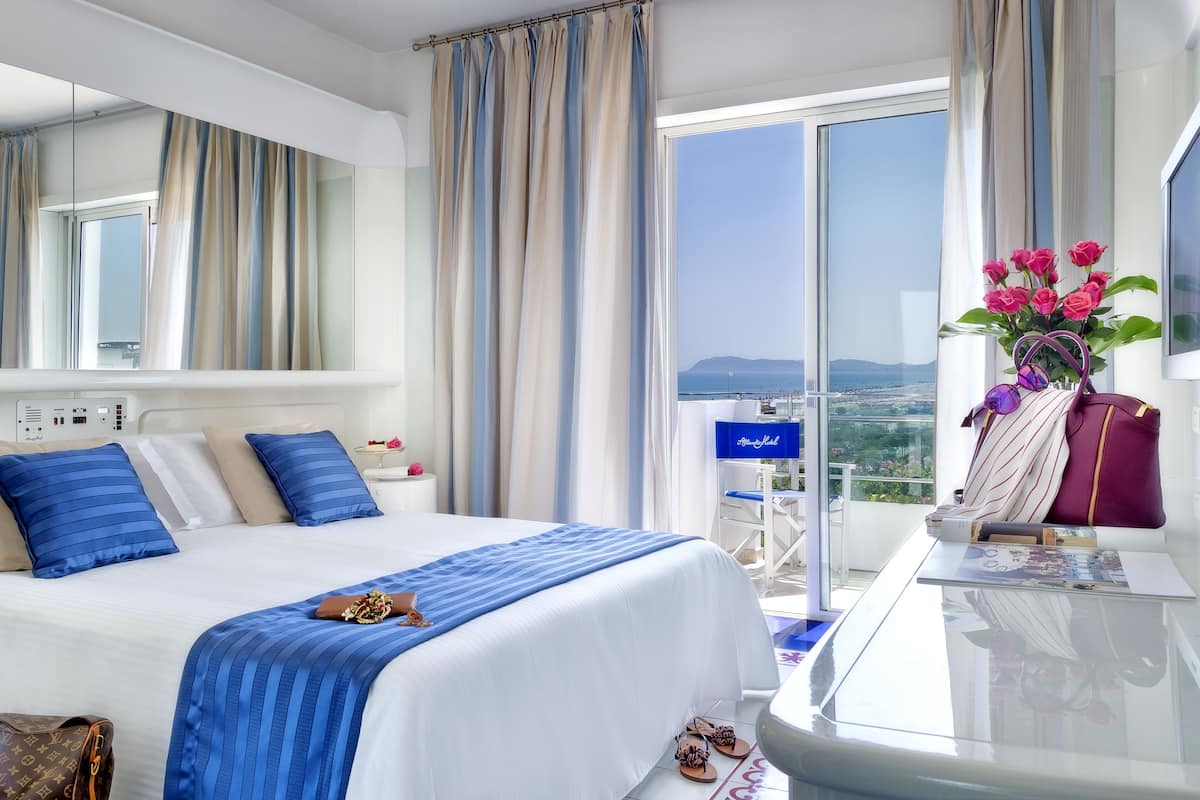 A hotel room with a large bed, a desk with a laptop, a view of the sea, and a balcony with flowers.