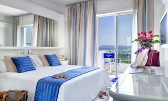 Habitación de hotel con una cama grande, un escritorio con una computadora portátil, vistas al mar y un balcón con flores.