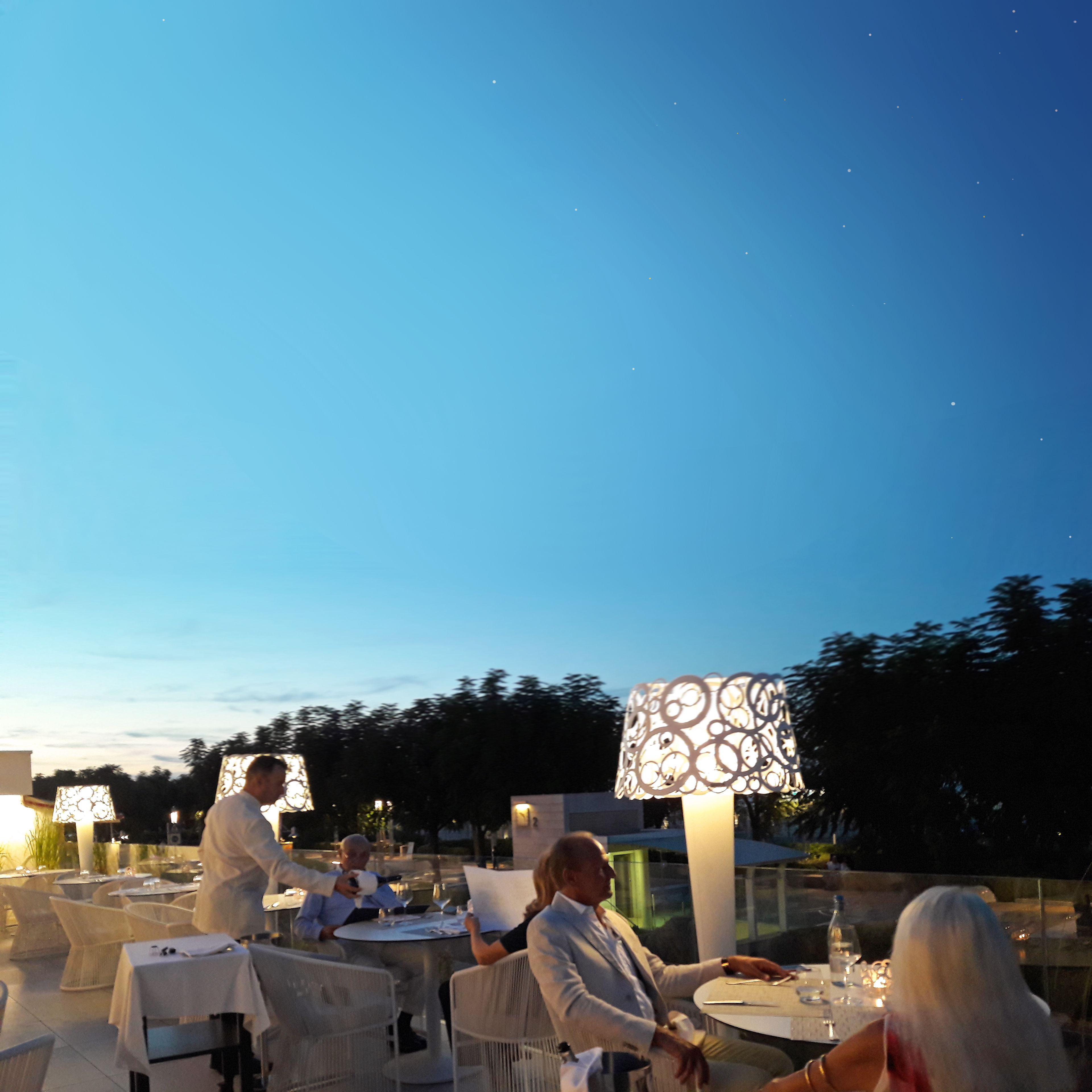 Un área de comedor al aire libre con mesas y sillas, iluminada por lámparas decorativas.