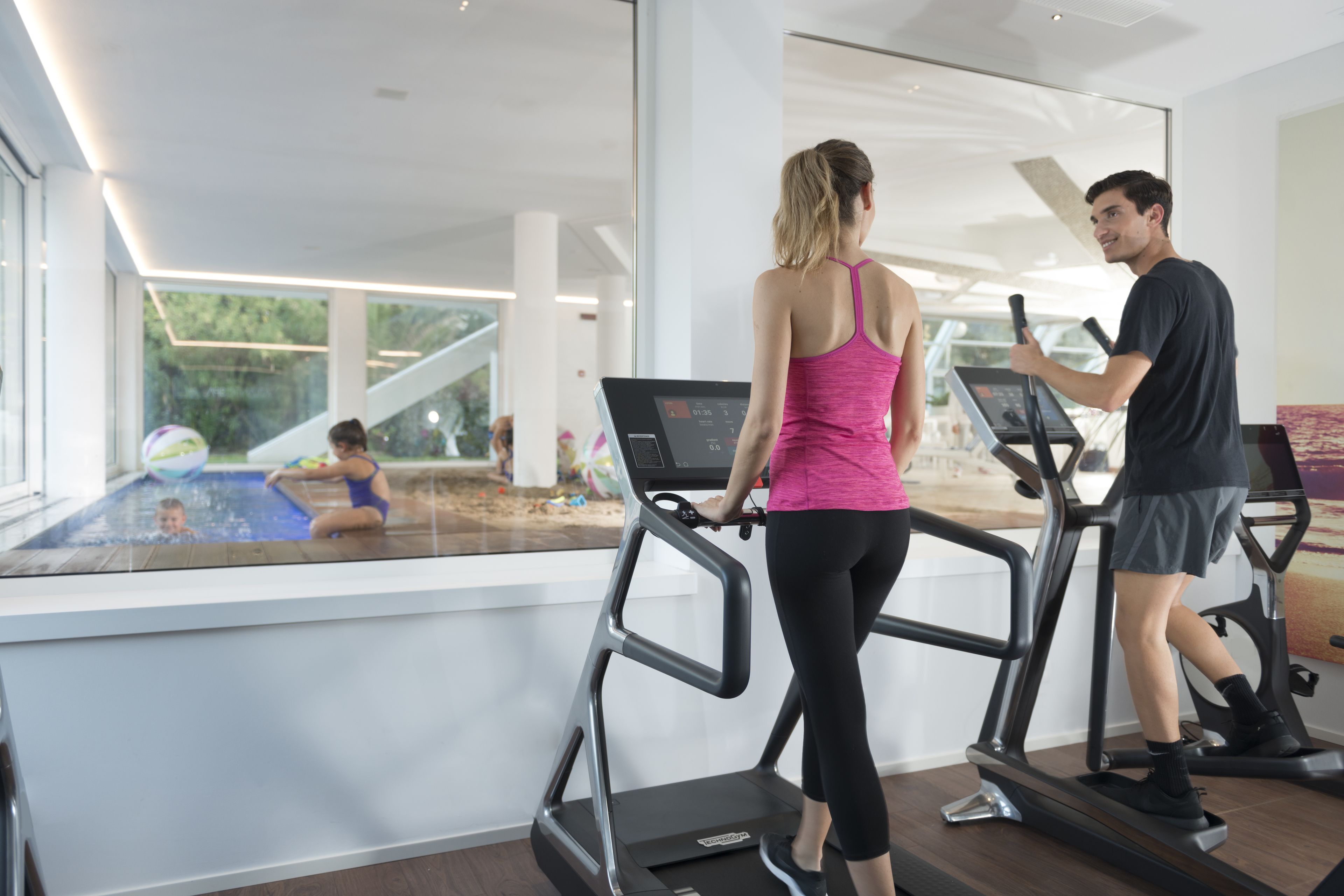 Un gimnasio con cintas de correr, una zona de piscina y grandes ventanales.