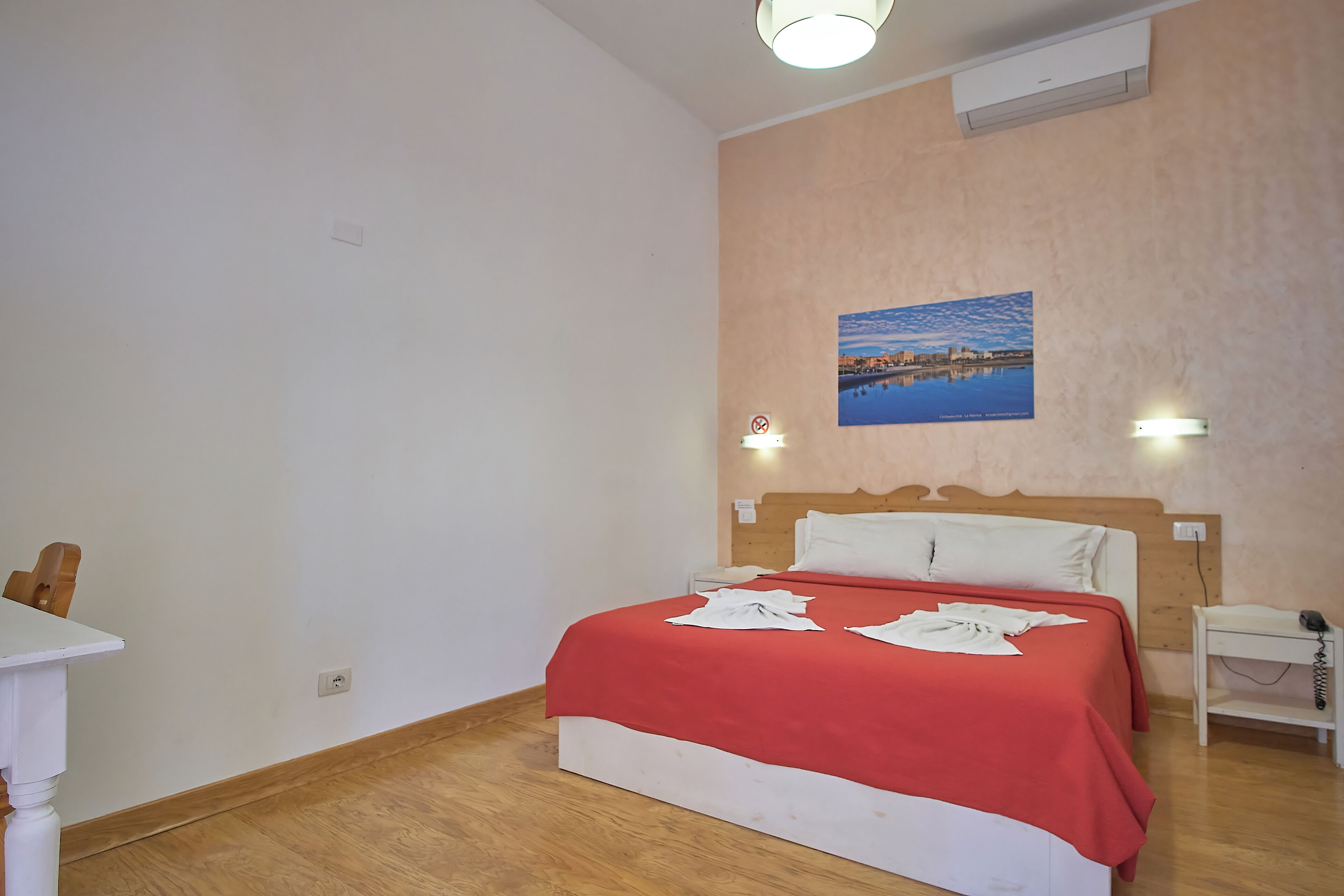 A bedroom with a wooden floor, a bed with a red blanket, a nightstand, a wall-mounted light, and a painting of a landscape.