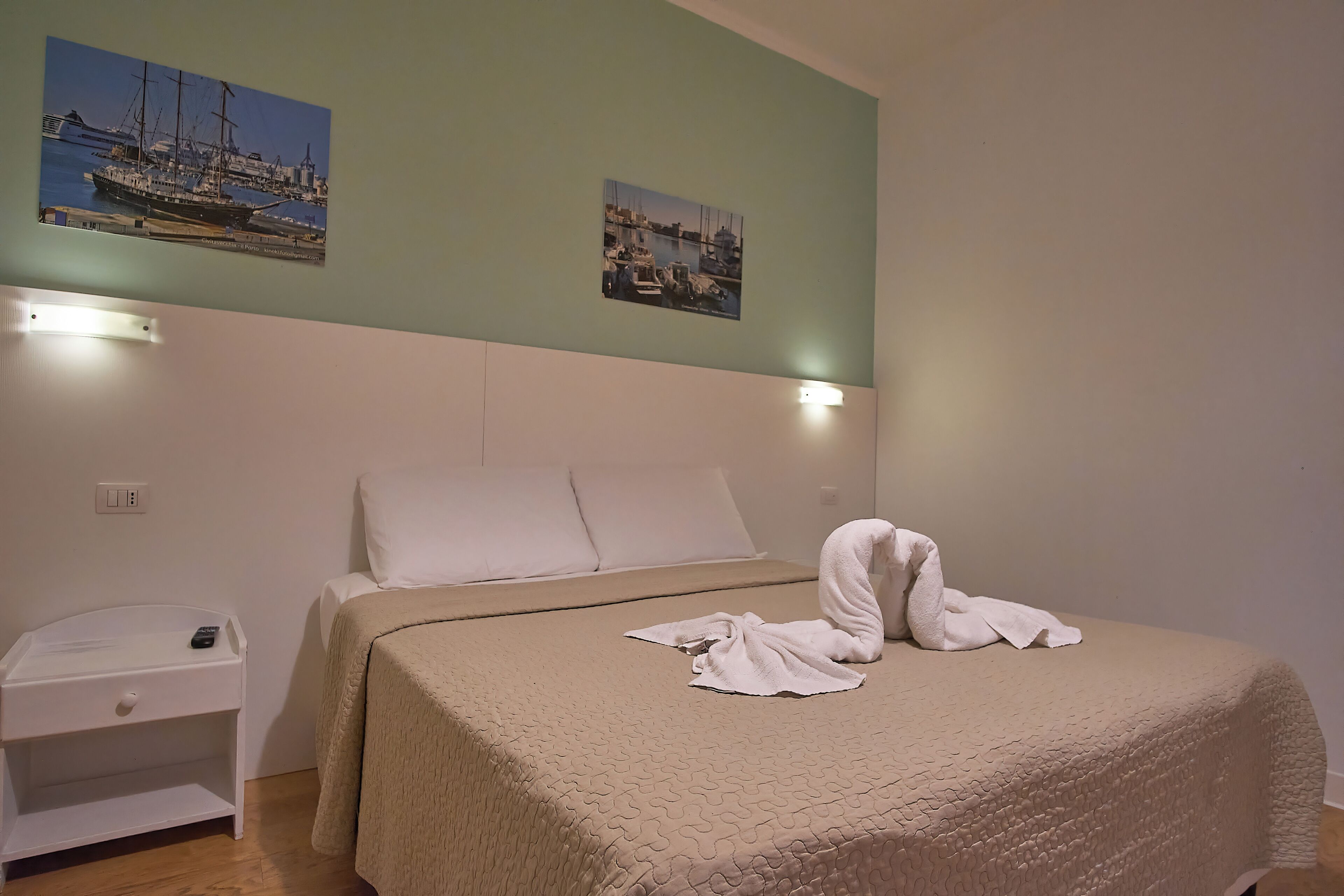 Ein ordentlich bezogenes Bett mit einer beigen Steudecke, ein weißer Nachttisch mit Fernbedienung und zwei gerahmte Bilder einer Marina an der Wand.