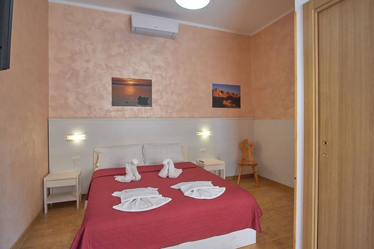 A hotel room with a red bedspread, two wall-mounted lights, a wooden chair, and two framed pictures on the wall.