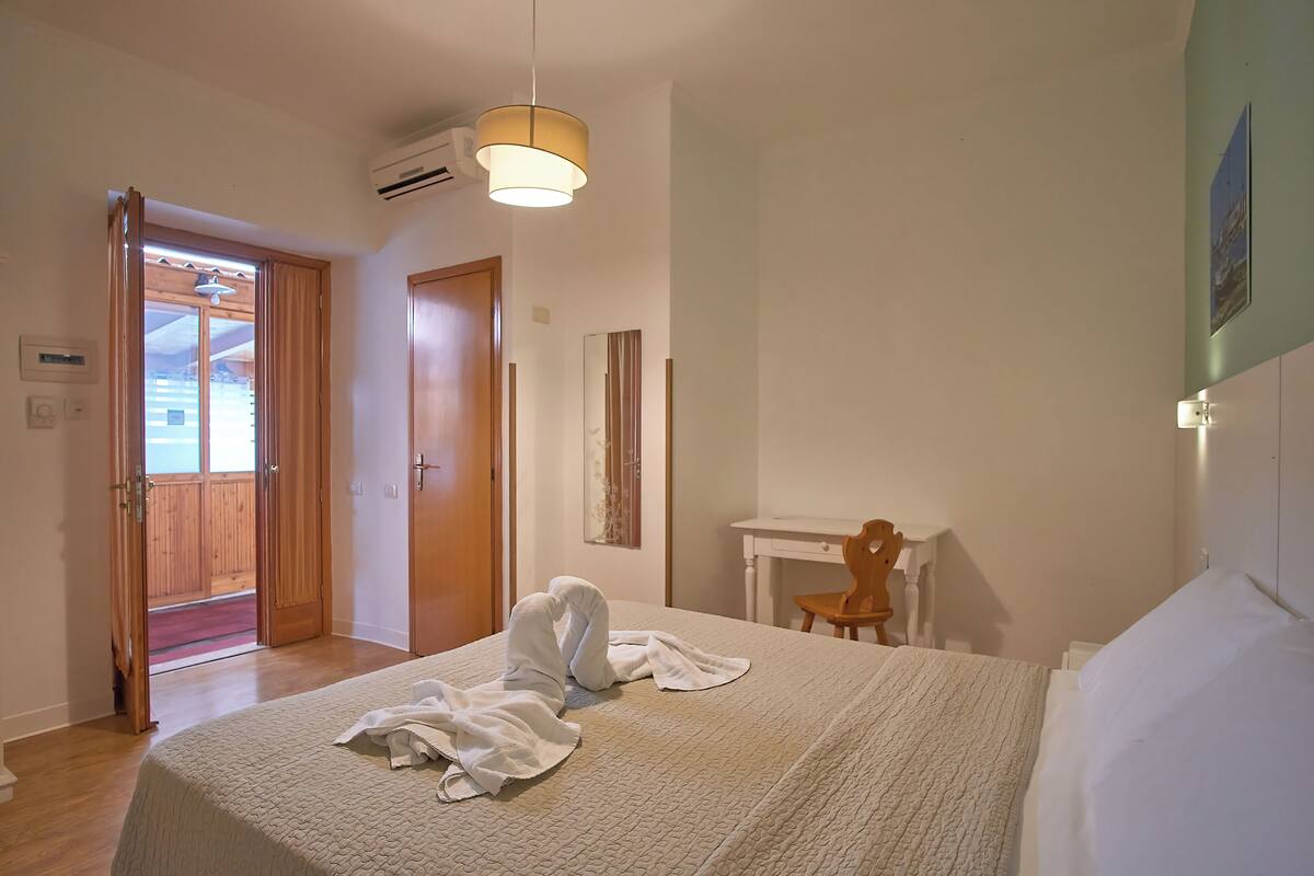 A bedroom with a bed, a desk, and a chair. There is a wooden door leading to an outdoor area.