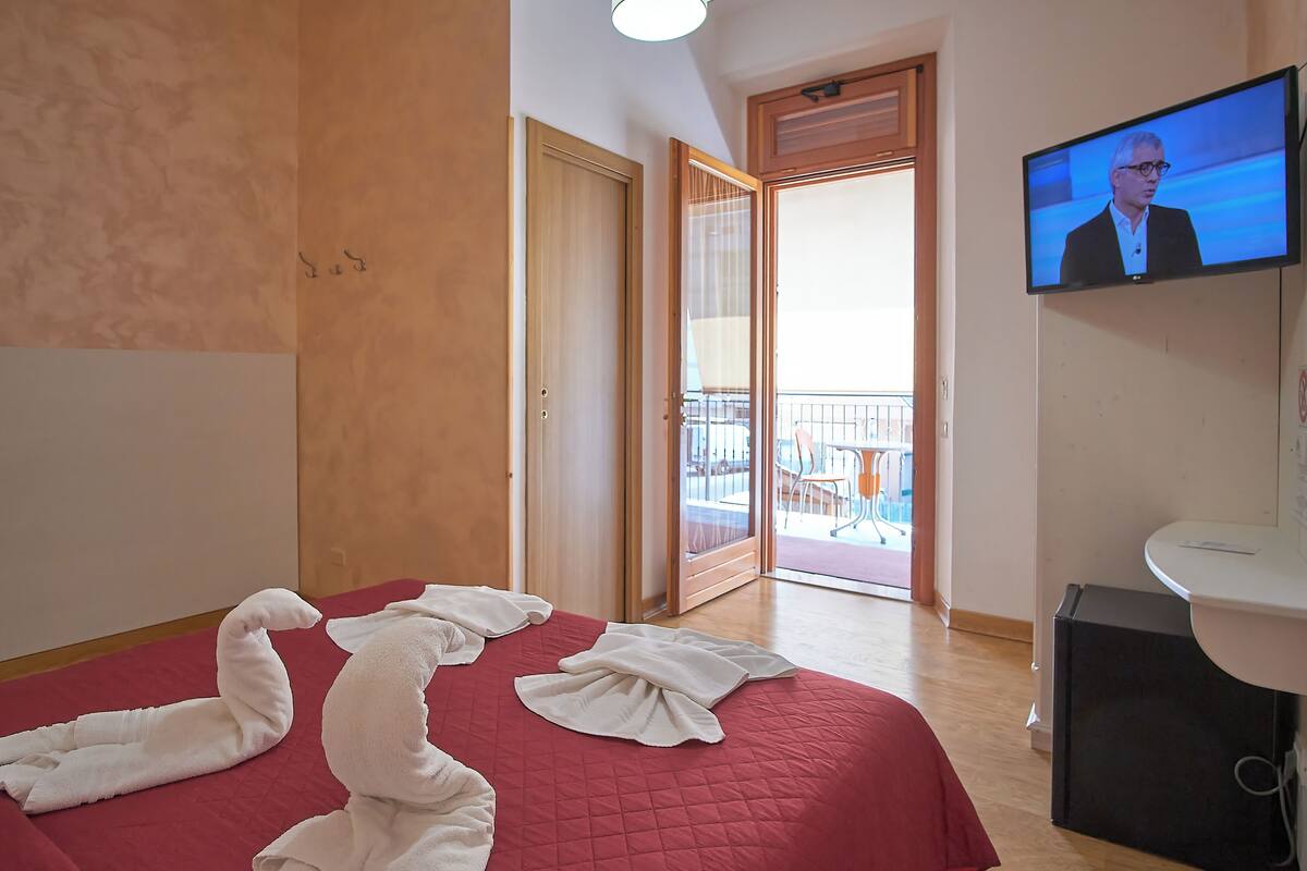 A hotel room with a red bedspread, a television mounted on the wall, and a balcony with a view of the outside.