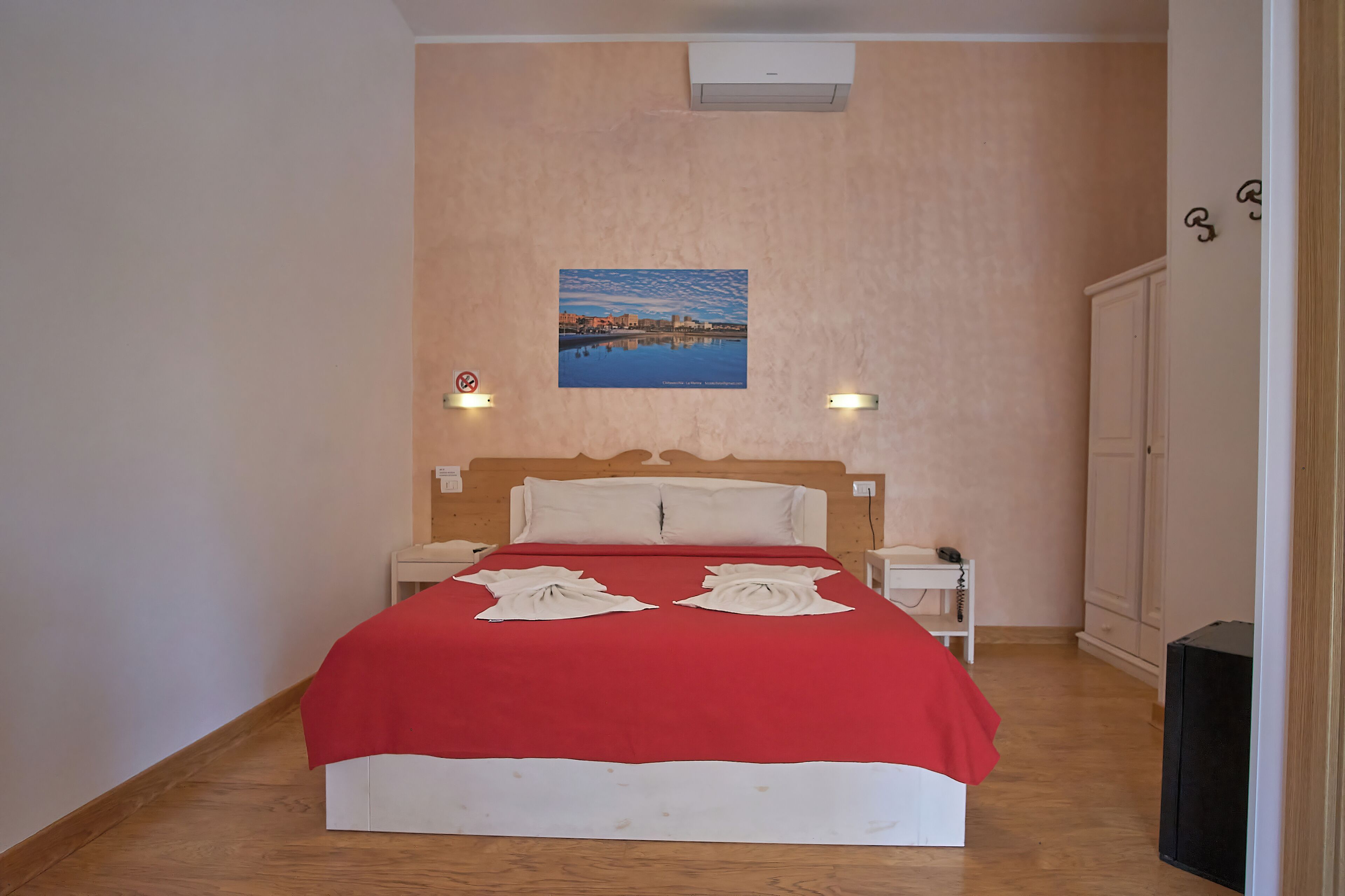 Un letto rifatto con coperta rossa e cuscini bianchi, un comodino con un telefono, un condizionatore d'aria a parete e un quadro che raffigura un paesaggio urbano.