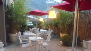 Un patio al aire libre con mesas y sillas, rodeado de plantas en macetas y sombrillas.