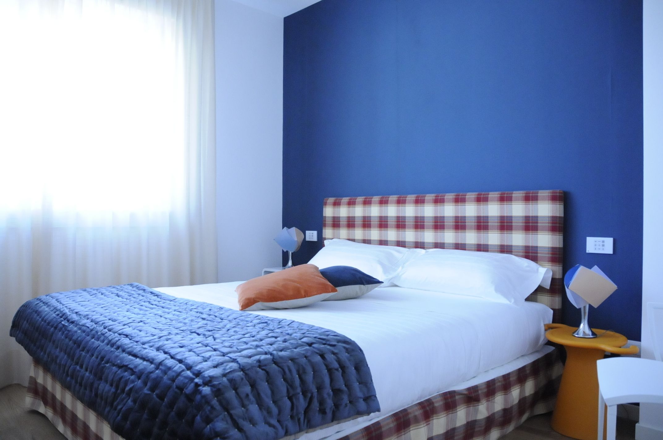 Ein ordentlich bezogenes Bett mit kariertem Kopfteil, einer blauen Decke und einem weißen Kopfkissen. Ein kleiner Nachttisch mit Lampe und Stuhl sind ebenfalls vorhanden.