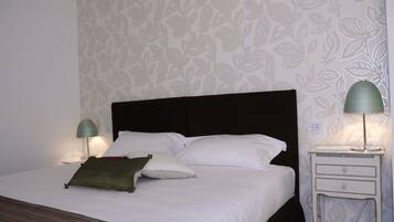 Una cama bien hecha con sábanas blancas y un almohadón verde, una mesita de noche con una lámpara, y una pared con estampado floral.