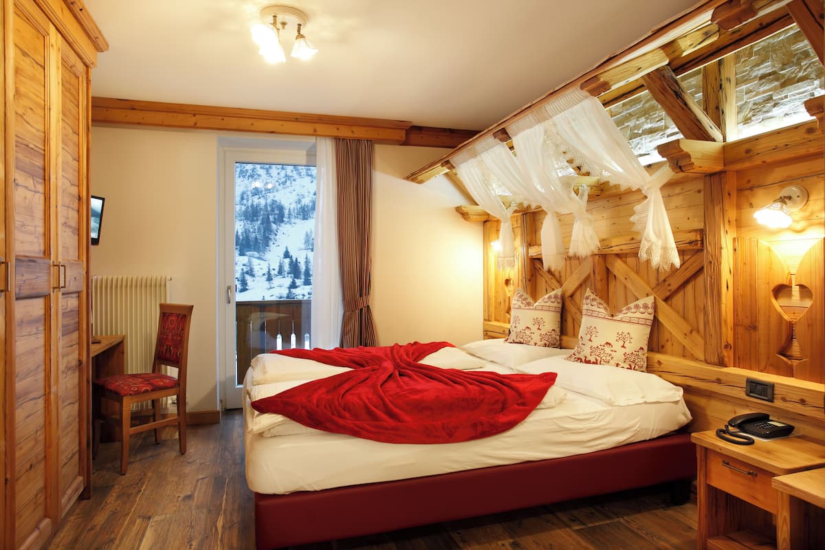 A wooden cabin bedroom with a bed, a chair, a window with a view of snow-covered trees, and a wooden ceiling with a light fixture.