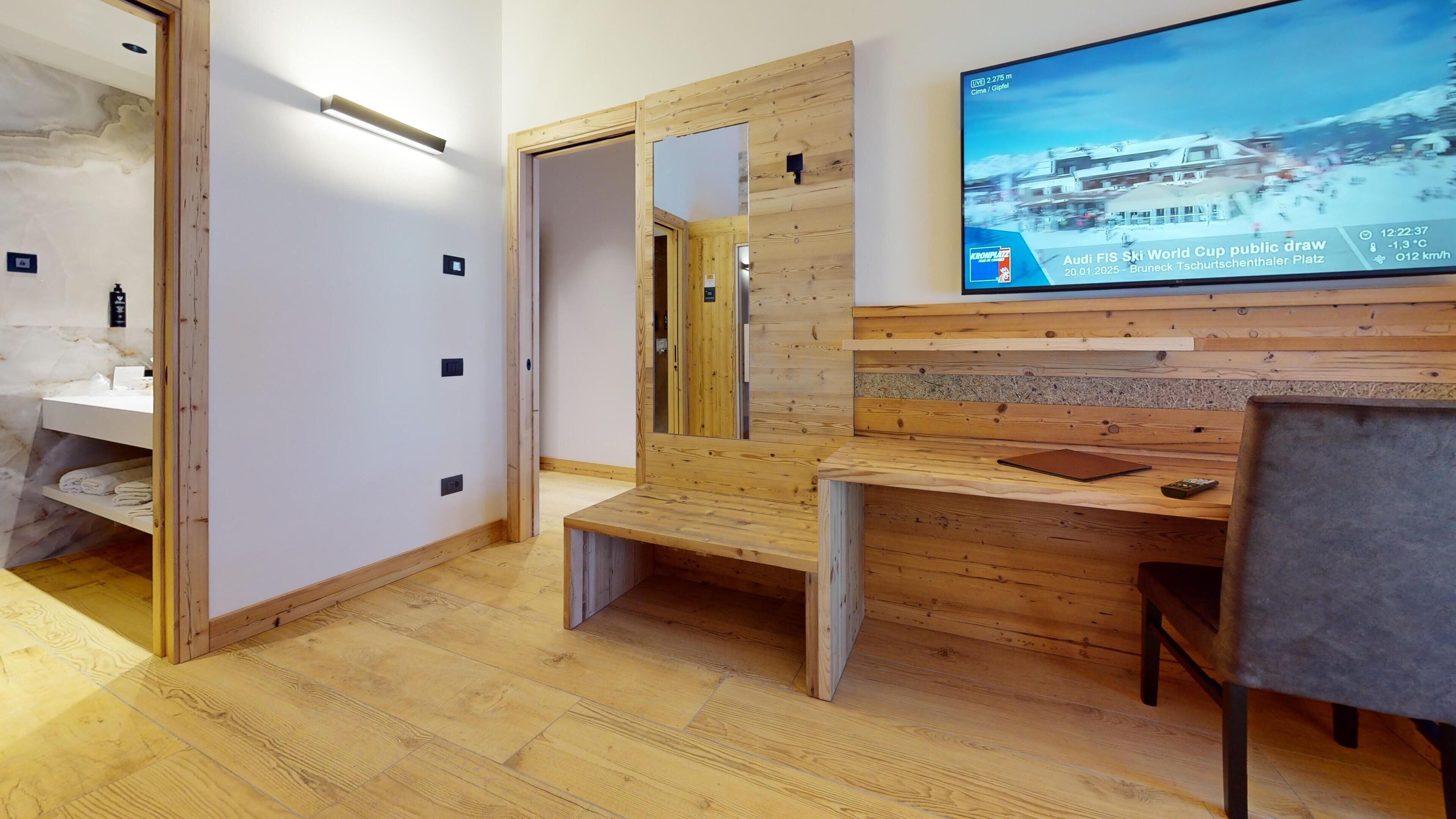A modern bathroom with wooden accents, a large mirror, and a television displaying a sports event.