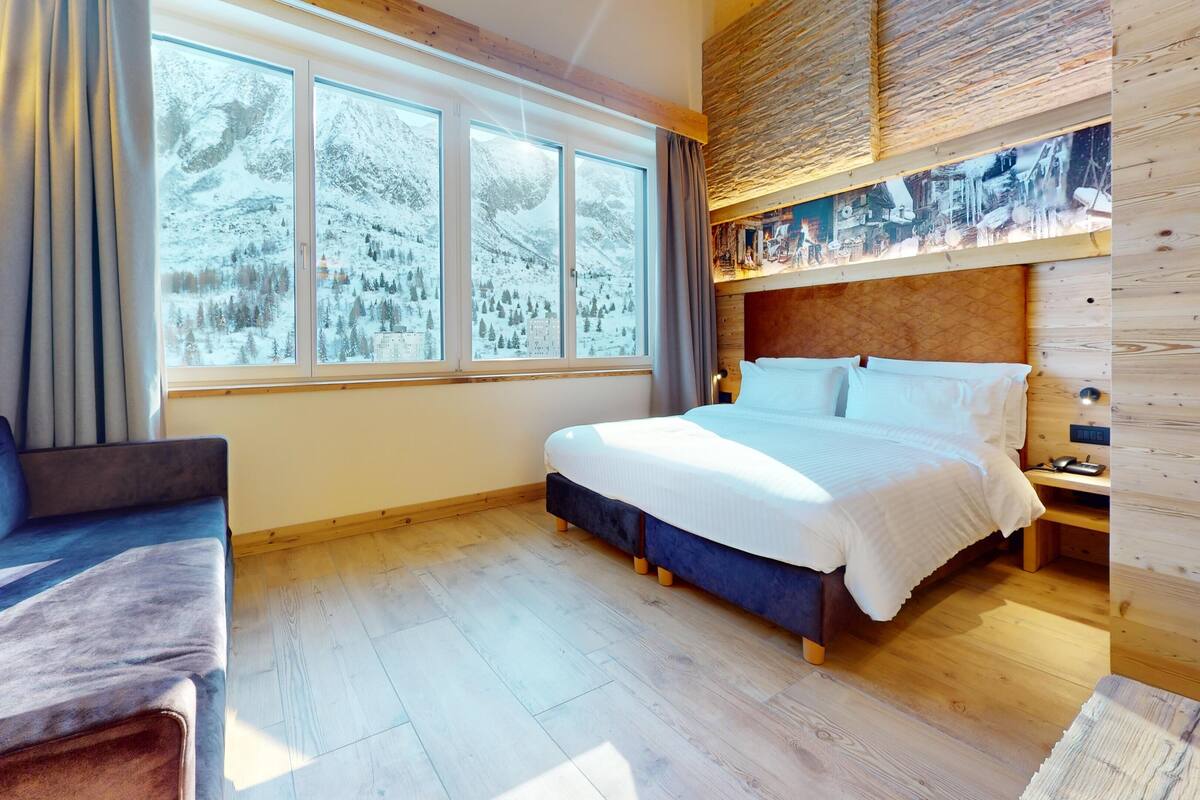 A cozy room with a large bed, a sofa, and a view of snowy mountains through large windows.