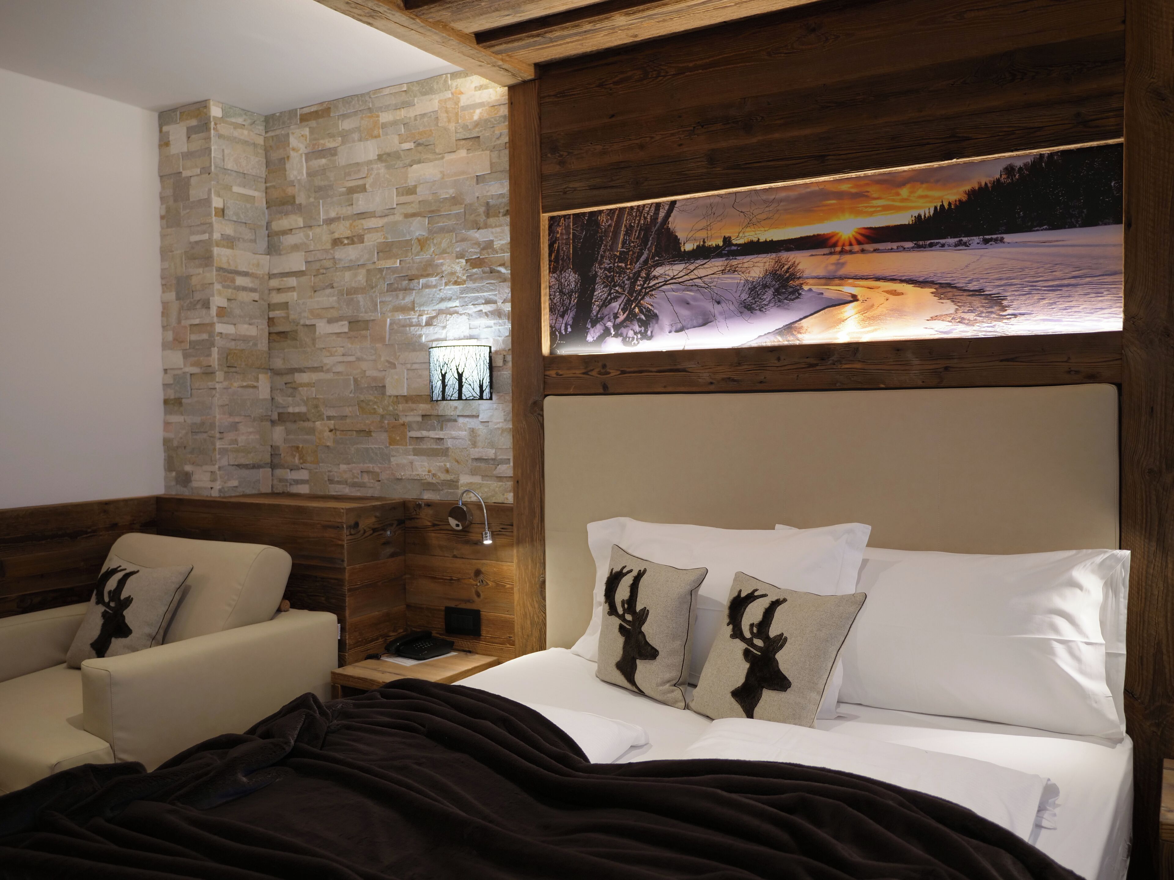 A cozy hotel room with a stone wall, a bed with white bedding and deer-themed pillows, a wooden headboard, and a painting of a winter landscape above the bed.