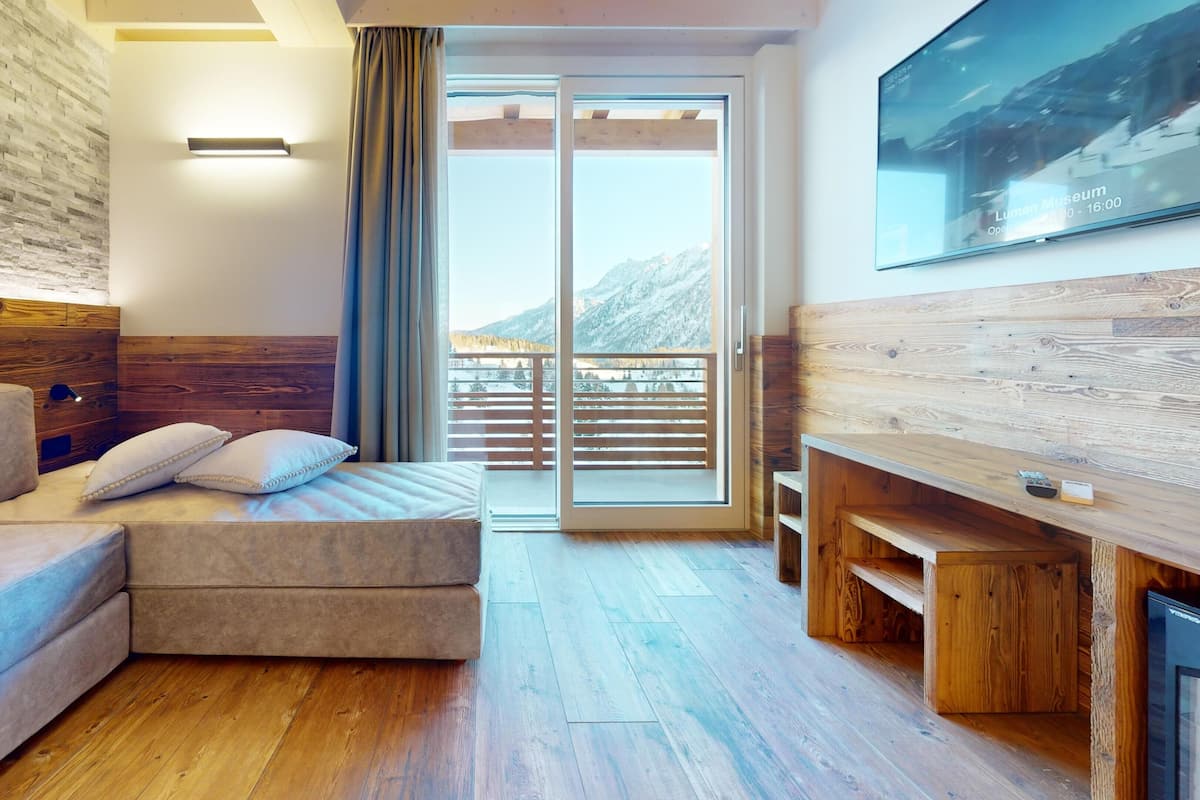A modern room with a wooden headboard, a sofa, a small table, and a sliding glass door leading to a balcony with a mountain view.