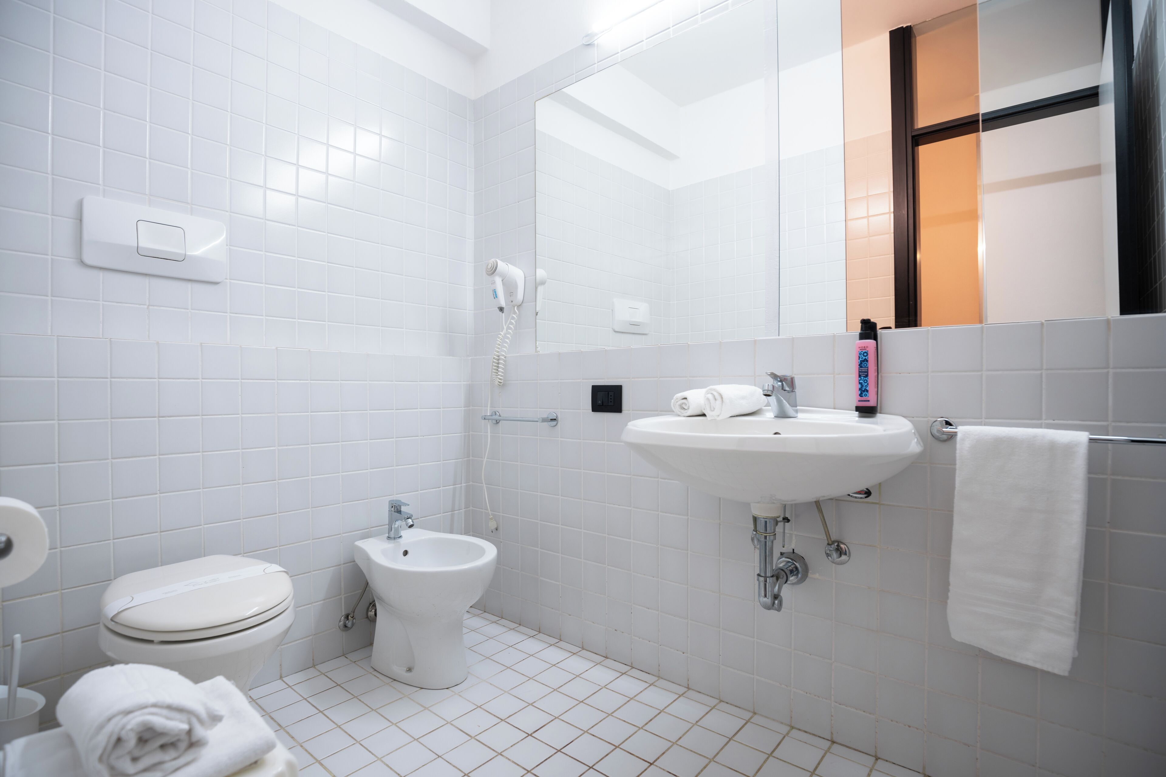A modern bathroom with white tiles, a toilet, bidet, sink, and a towel rack.
