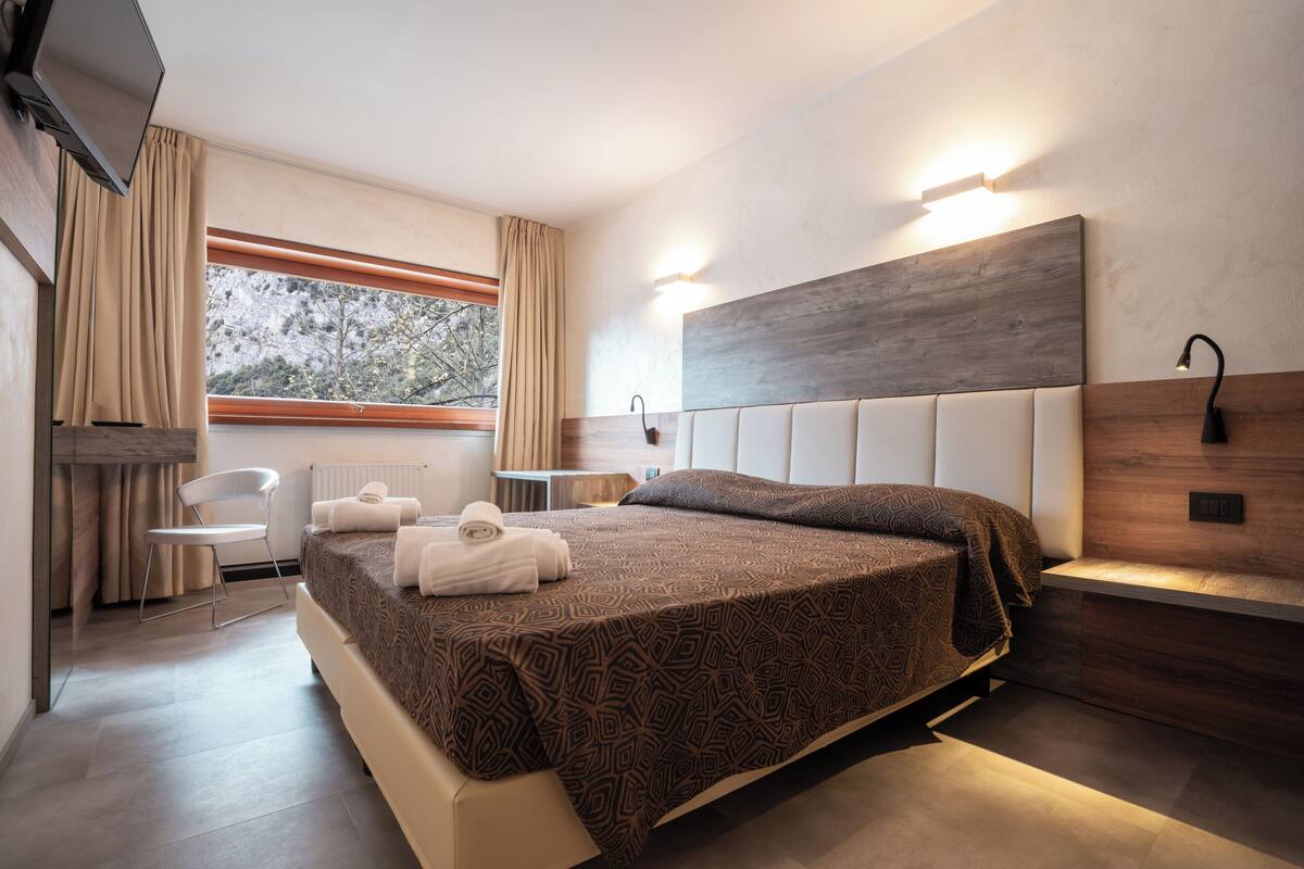 A modern hotel room with a large bed, a chair, a desk, and a view of trees through the window.