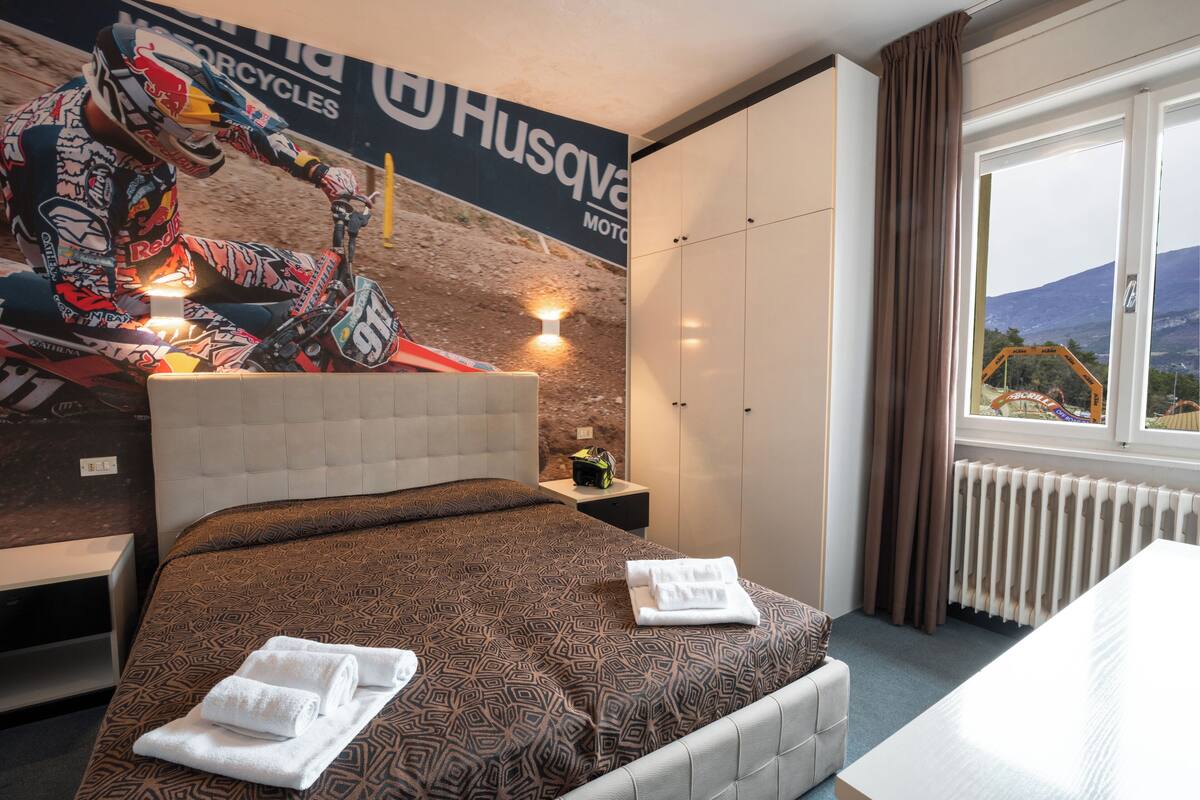 A hotel room with a bed, a nightstand, a window with a view of a mountain and construction site, and a wall with a motorcycle advertisement.
