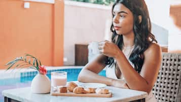 Une personne est assise à une table devant une tasse blanche, un verre de lait et des croissants. Sur la table, on aperçoit un petit vase blanc contenant une fleur rose.