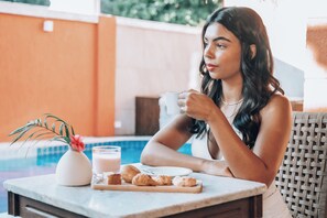 Una persona sentada a una mesa con una taza blanca, un vaso de leche y croissants. Hay un pequeño florero blanco con una flor rosa sobre la mesa.