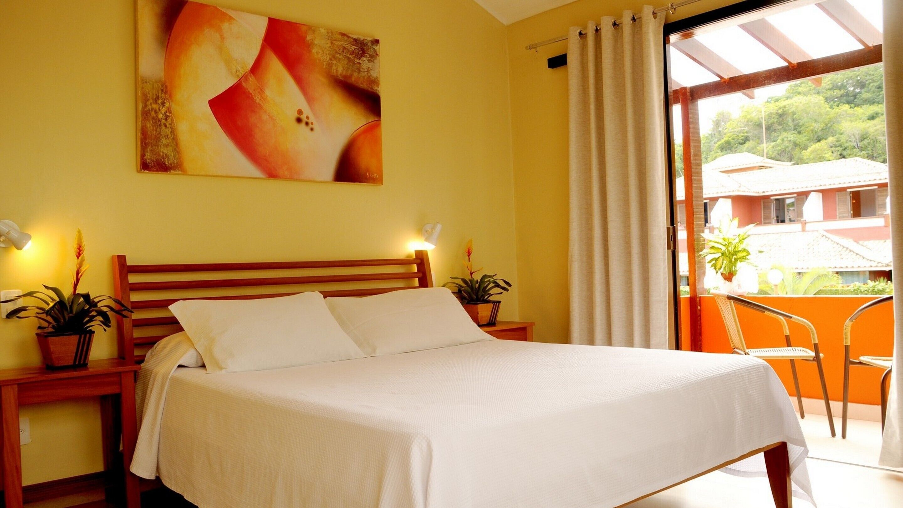 A bedroom with a wooden headboard, a large bed, two bedside tables with lamps, and a view of a balcony with a chair and potted plants.
