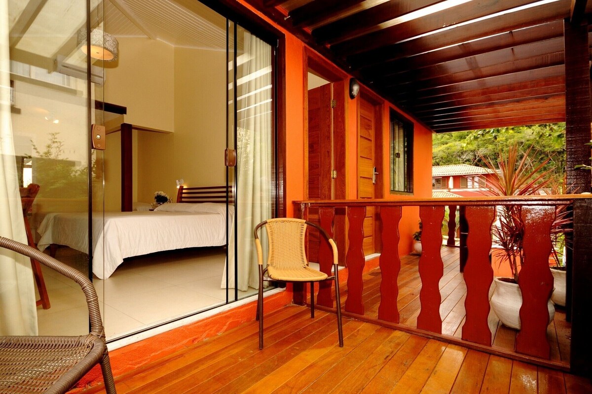 A room with a bed, a wicker chair, and a balcony with a wooden railing.