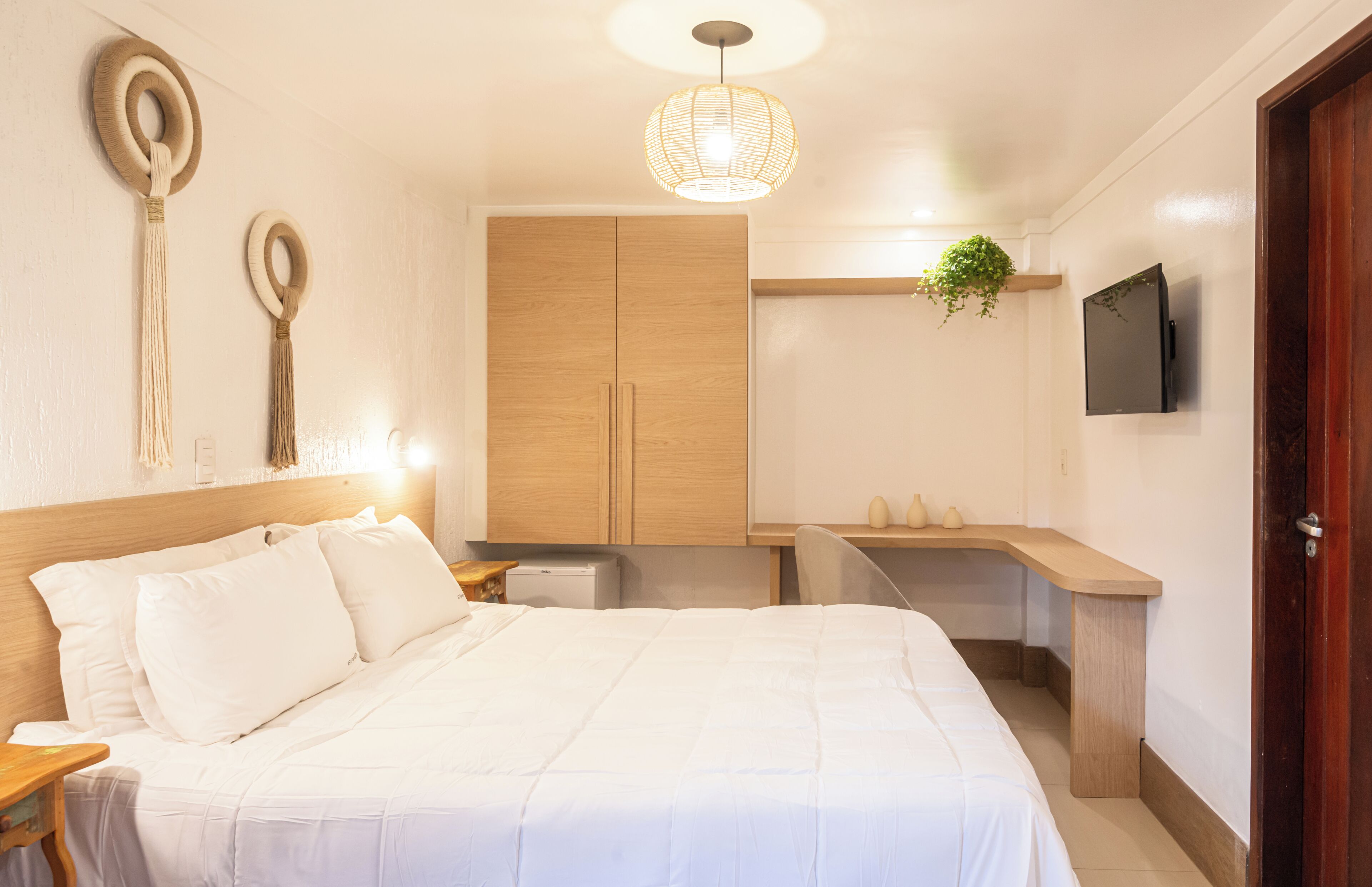 Un dormitorio con cama, armario de madera, mesita de noche, planta colgante y televisor en la pared.