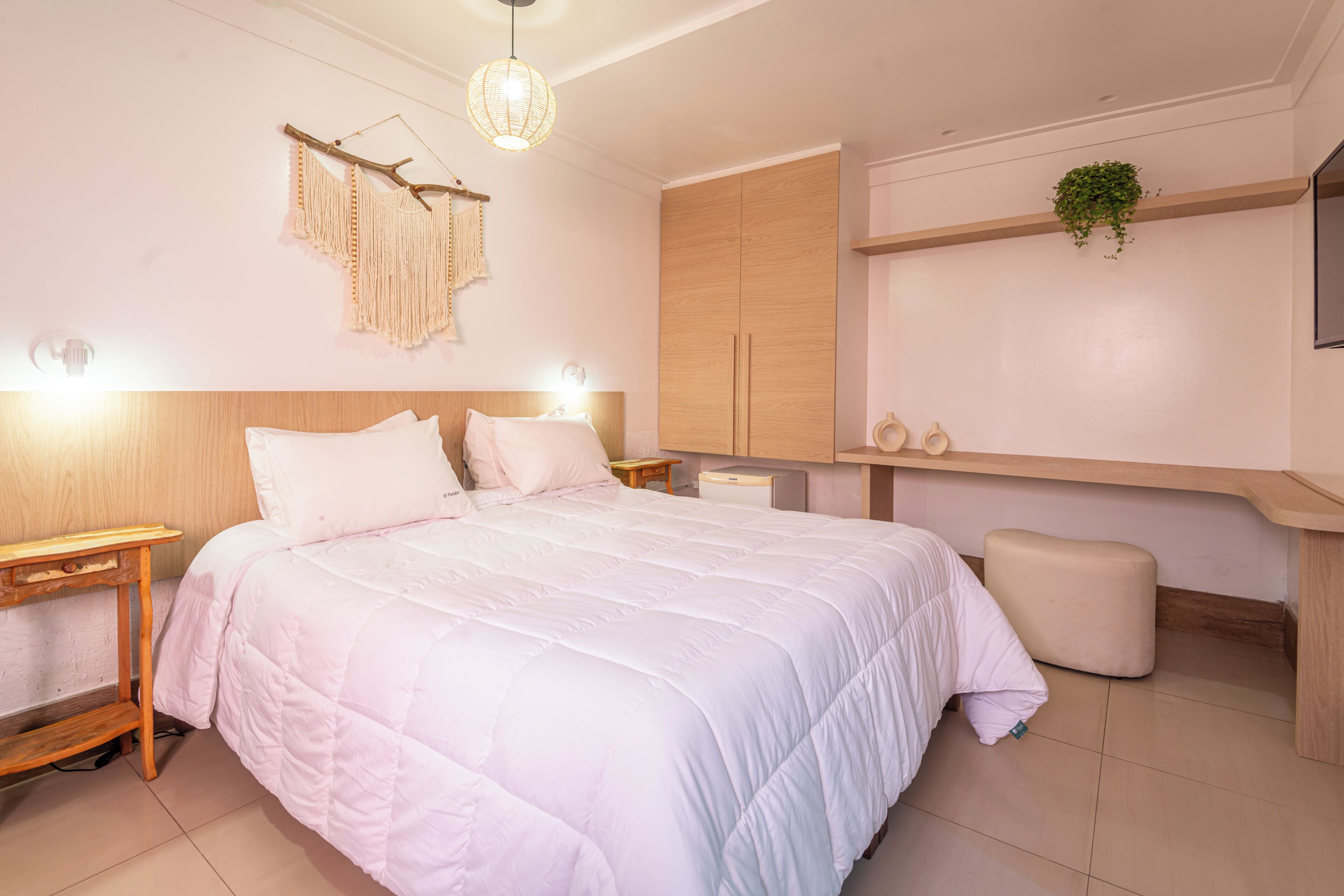 A bedroom with a large bed, bedside tables, a wardrobe, a small stool, a hanging light fixture, and a plant on the wall.