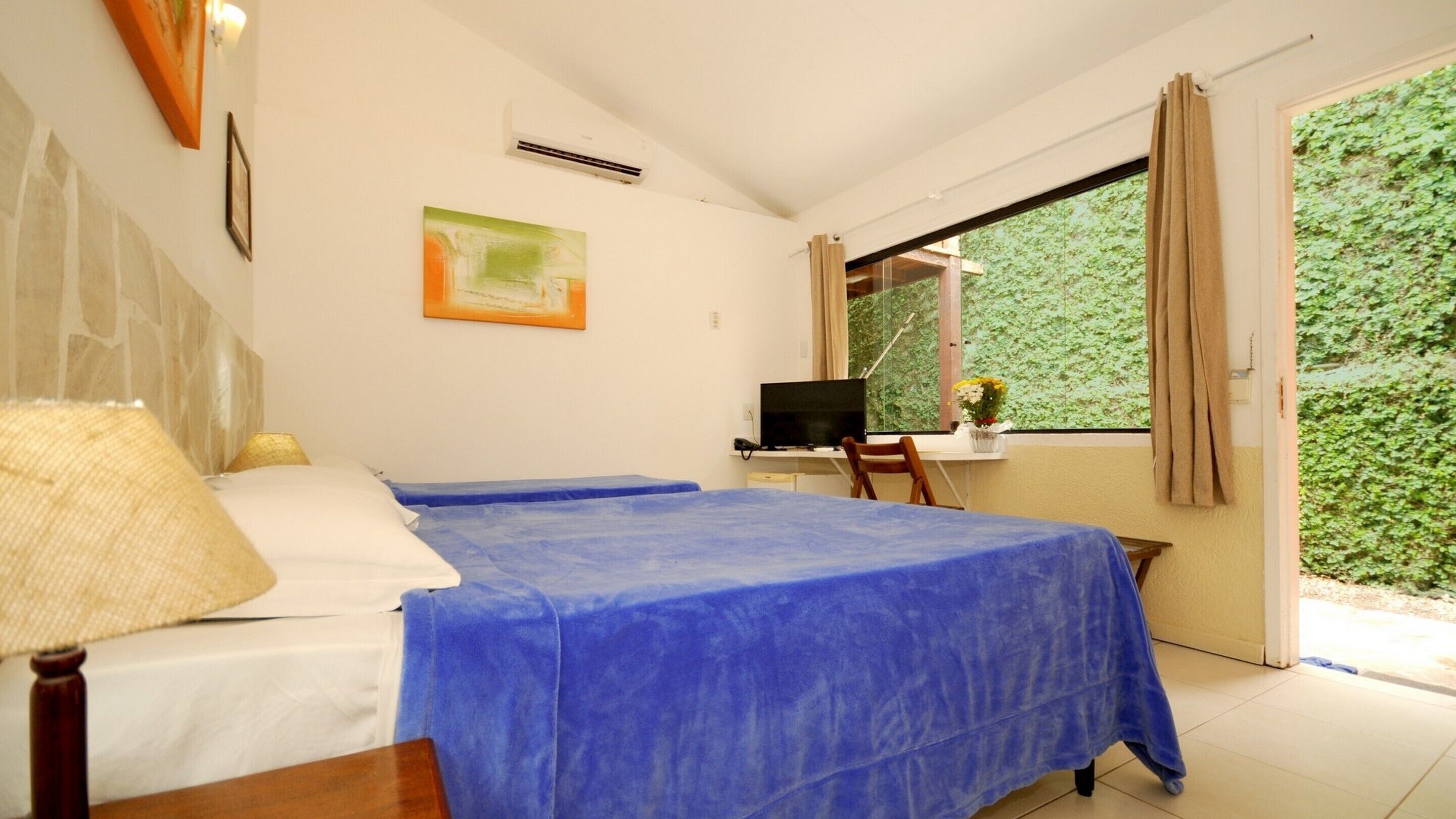 A bedroom with two beds, a desk with a computer, a window with a view of greenery, and a painting on the wall.