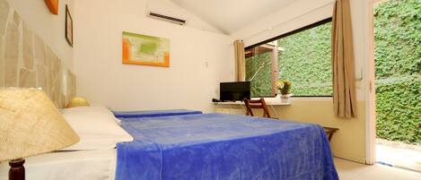 A bedroom with two beds, a desk with a computer, a window with a view of greenery, and a painting on the wall.