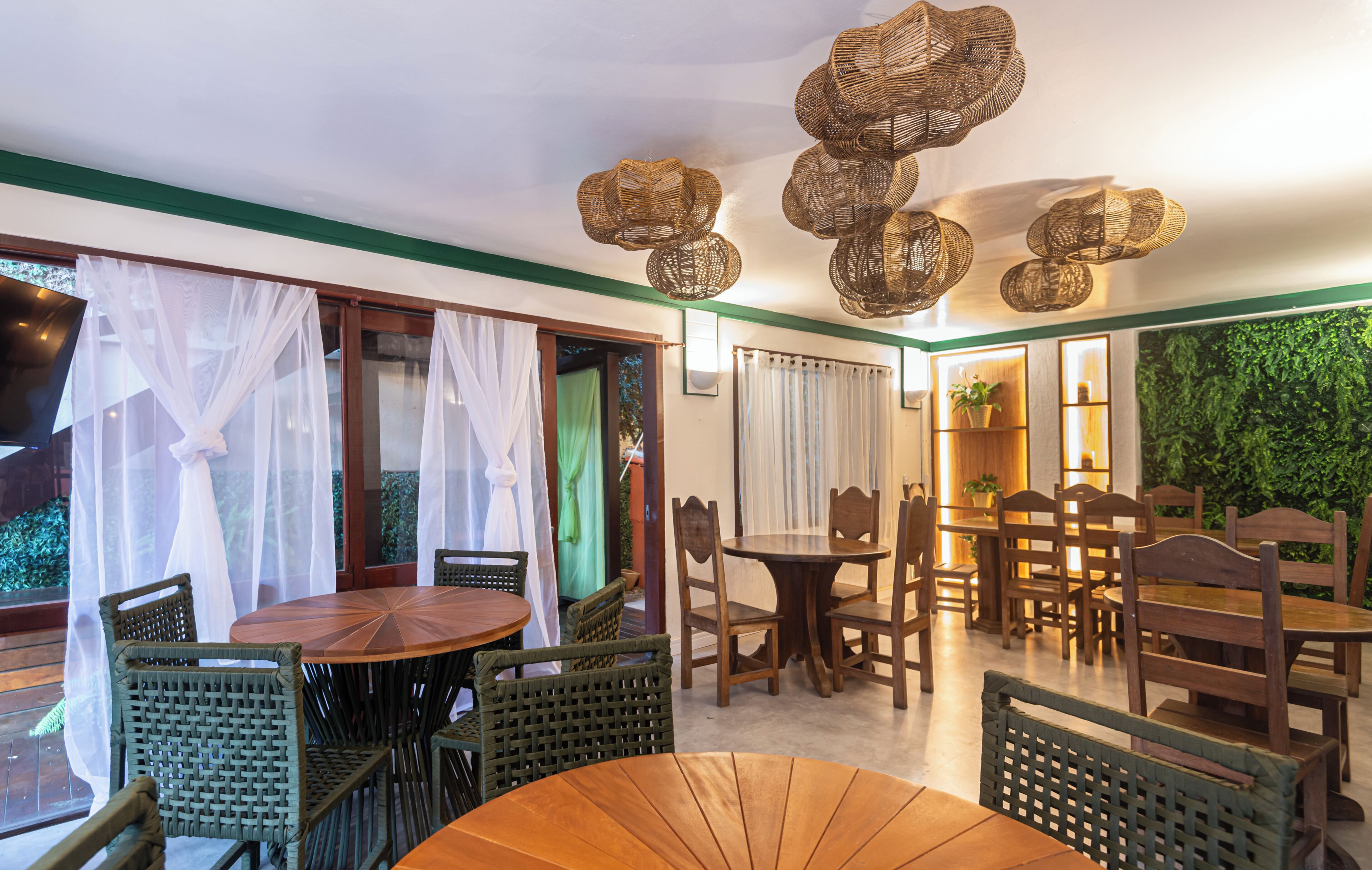 A restaurant interior with round tables and chairs, a green wall with a plant, and a ceiling with woven light fixtures.