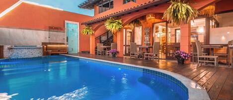 Una piscina con cubierta de madera, asientos exteriores y un edificio con entrada de azulejos.