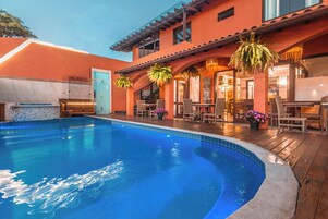 Una piscina con cubierta de madera, asientos exteriores y un edificio con entrada de azulejos.