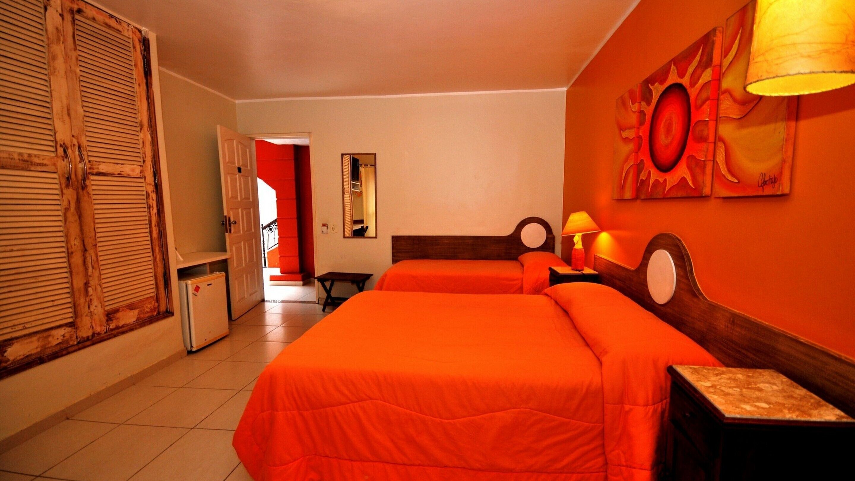 Habitación de hotel con dos camas, una mesita, una lámpara y una puerta que da a otra habitación.
