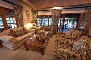 A cozy living room with two sofas, a coffee table, and a lamp. There are large windows with curtains, and a view of the outdoors. - Blyde River Canyon Lodge (Hoedspruit)