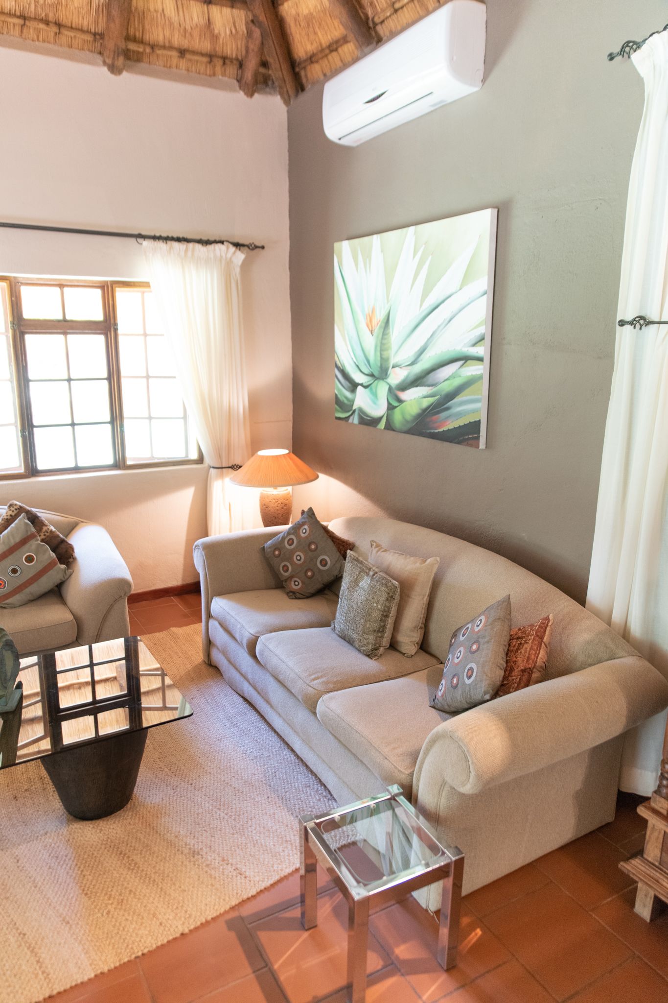 A living room with a beige sofa, a glass coffee table, a lamp, and a large plant on the wall.