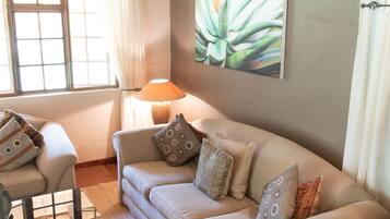 A living room with a beige sofa, a glass coffee table, a lamp, and a large plant on the wall.