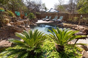 Quintal com piscina, cercado por pedras e plantas. Há espreguiçadeiras e um cercado de bambu.