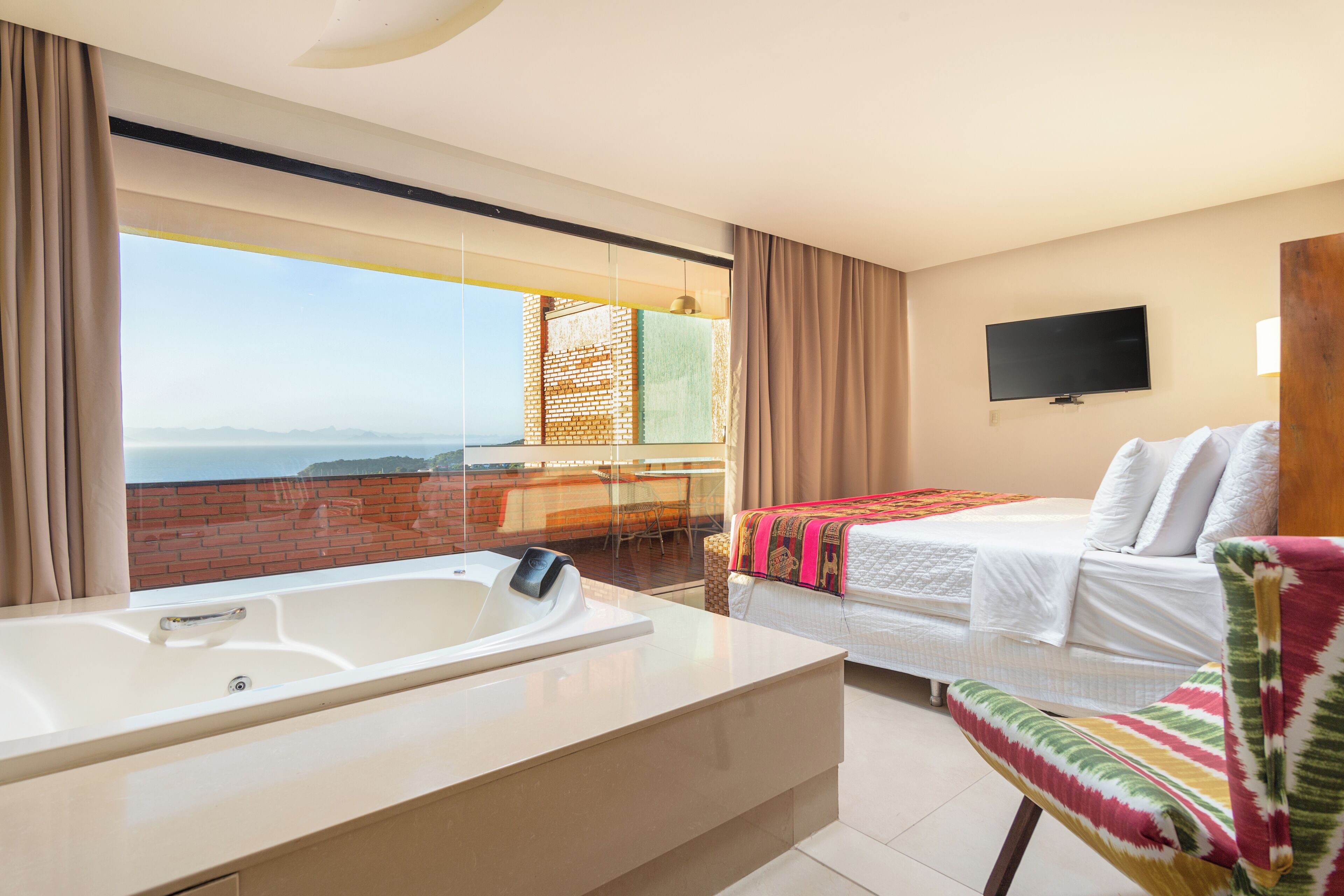 A hotel room with a large window, a bed, a chair, a TV, and a bathtub.
