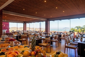 A spacious dining area with tables and chairs, a buffet with various food items, and large windows offering a view of the ocean. - Costa do Sol Boutique Hotel (Búzios)