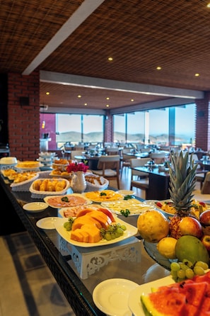 A buffet with various fruits and pastries, a brick wall, and a view of the mountains. - Costa do Sol Boutique Hotel (Búzios)