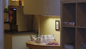 Une petite cuisine équipée d’une machine à café, une petite table avec des tasses et un panier de viennoiseries, ainsi qu’une bibliothèque avec des journaux.