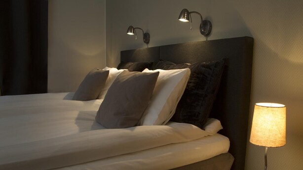 Un lit bien fait, avec du linge de lit blanc et plusieurs oreillers, une lampe de chevet et deux lampes de lecture fixées au mur.