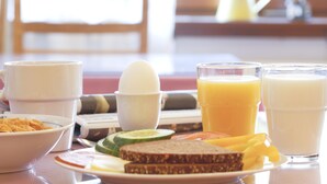 Une table de petit-déjeuner avec un bol de céréales, une tasse de café, un verre de jus d’orange, un verre de lait, une assiette de pain grillé, des tranches de concombre et une tomate.
