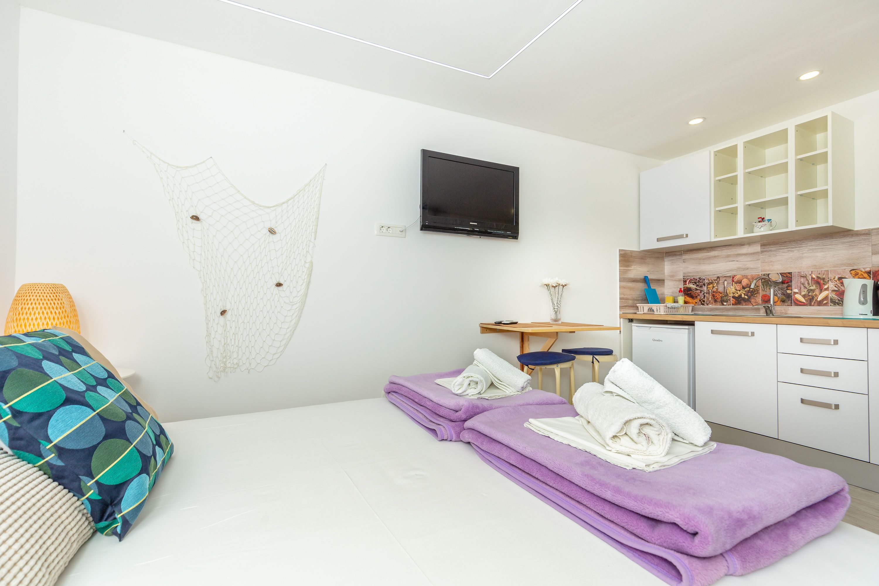 A modern hotel room with a bed, a desk, a television, and a kitchenette.