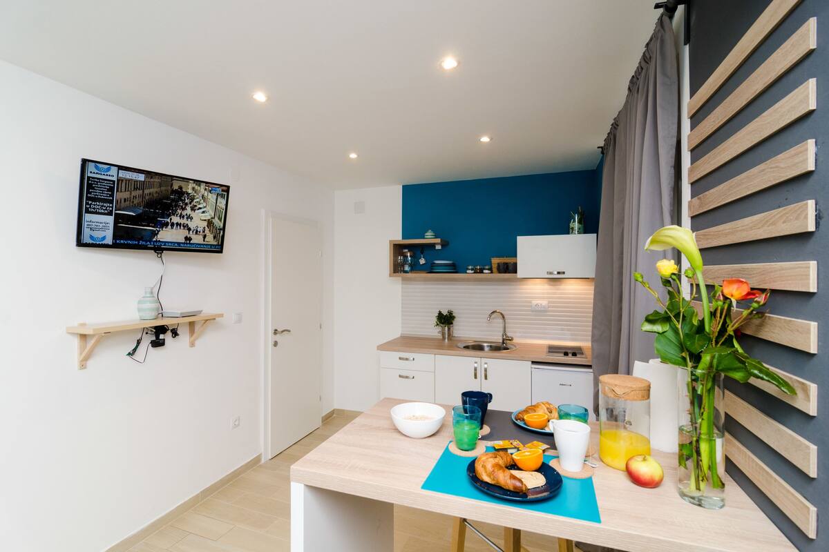 A modern kitchen with a blue accent wall, a dining area with a table set for breakfast, and a mounted television on the wall.