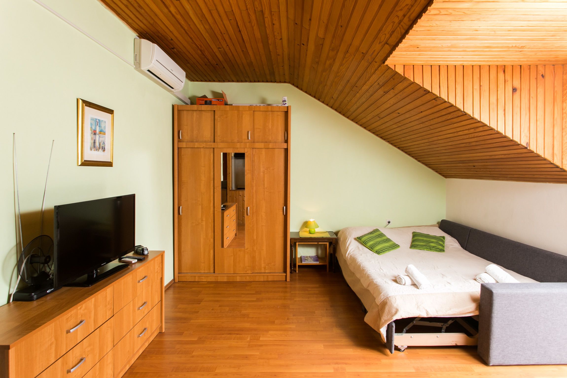 Une chambre avec un plafond en bois, une armoire, un lit avec des oreillers, une table de chevet et un téléviseur posé sur une commode en bois.