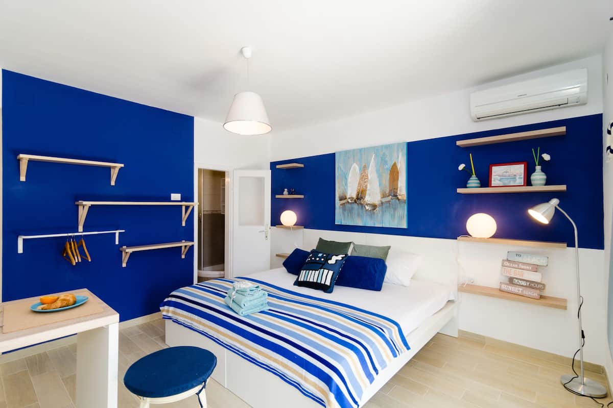 A bedroom with a bed, bedside tables, a desk, and a chair. There are shelves with decorative items and an air conditioning unit on the wall.