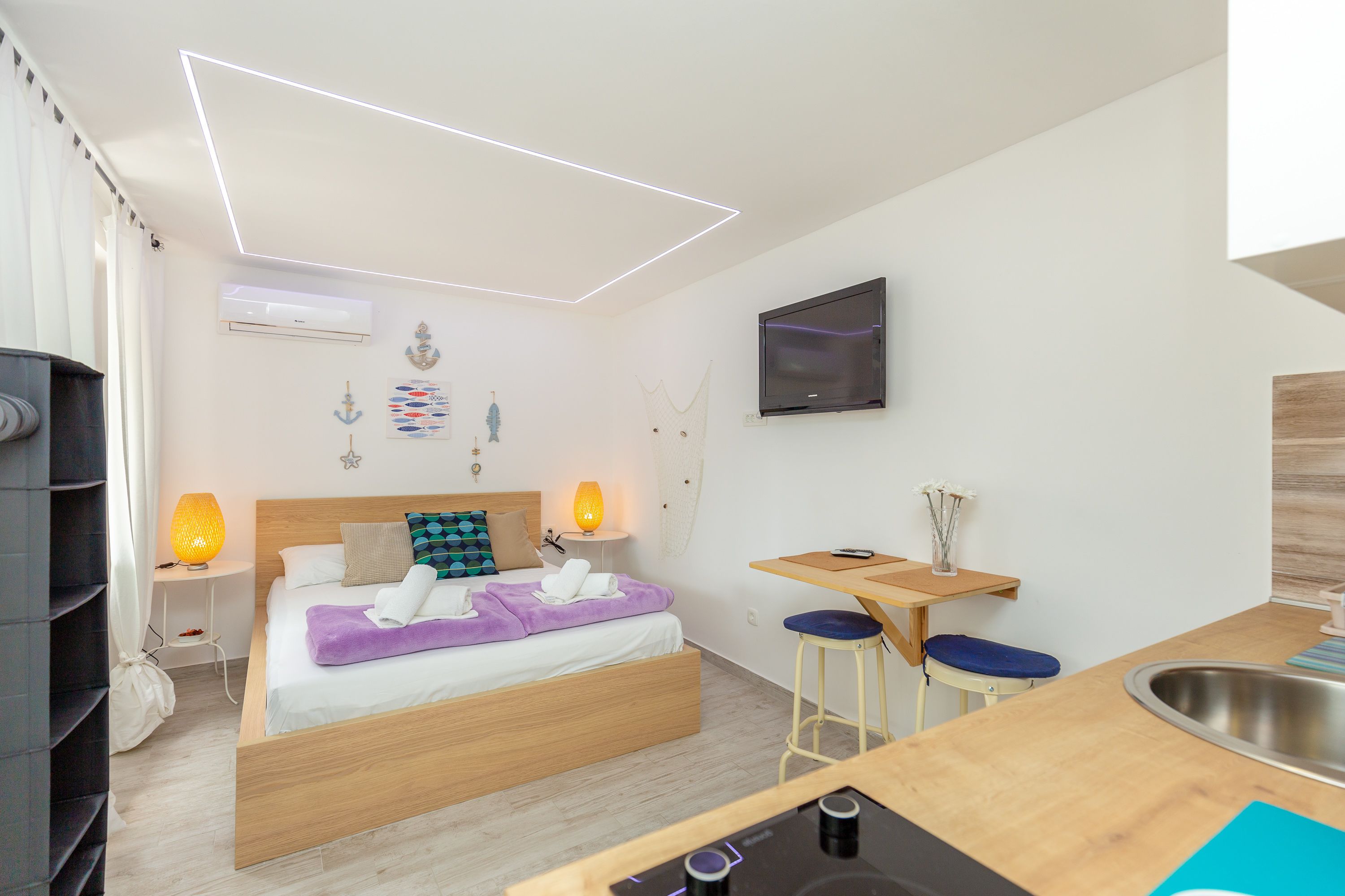 A compact bedroom with a bed, bedside lamps, a TV, a small table, and a sink.